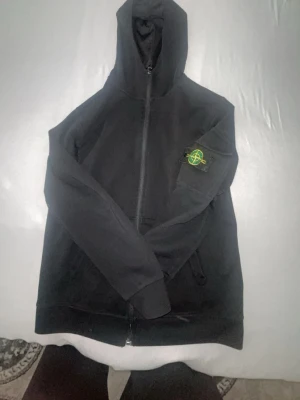 Svart hoodie från Stone Island - Svart hoodie med dragkedja från Stone Island. Klassisk modell med huva och ribbade muddar. Ikonisk logotyp-patch på vänster ärm. Perfekt för en avslappnad streetwear-look. Tillverkad i mjukt material för extra komfort.