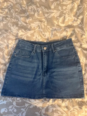 Blå jeanskjol med fickor - Snygg blå jeanskjol med klassisk femficksdesign och hällor för skärp. Jeanskjolen är aldrig använd, alltså helt ny.  Kjolen har en kort passform och stängs med knapp och dragkedja framtill. Perfekt att matcha med en t-shirt eller topp nu inför sommaren 