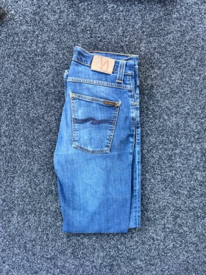 Blå slim fit jeans från Nudie Jeans - Snygga blå slim fit jeans från Nudie Jeans med klassisk femficksdesign och ljus tvätt. Jeansen har orangea sömmar, knappgylf och Nudie-logga på bakfickan. Perfekta för en avslappnad och trendig look. Liten lagning i skrevet men inget som syns, se sista bilden