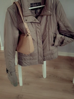 Beige pufferjacka med hög krage - Snygg beige pufferjacka med hög krage och dragkedja framtill. Från 2000 talet. Från MNG. Stl L men passar mig som har Xs-S. Jackan har quiltade sömmar och två fickor på sidorna. Ihopfällbar luva och fina stickade ändar på ärmarna. Perfekt för kalla dagar och ger en stilren look. Materialet är syntet och jackan har en klassisk, något kortare passform. 