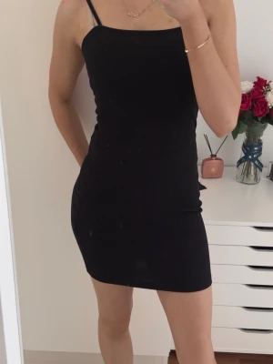 Svart tight kort klänning - Enkel och stilren svart klänning med smala axelband och tight passform. Klänningen är kort och har en minimalistisk design som passar perfekt för en trendig look. Materialet känns mjukt och följsamt mot kroppen.