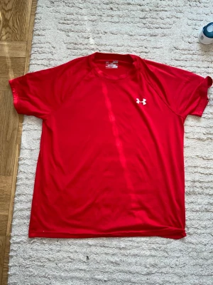Röd träningströja från Under Armour - Säljer en röd kortärmad träningströja från Under Armour med vit logga på bröstet. Tröjan är tillverkad i ett lätt och snabbtorkande syntetmaterial, perfekt för intensiva träningspass.