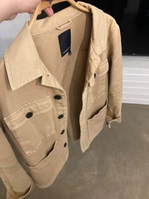 Beige overshirt från J.Lindeberg - Stilren beige overshirt från J.Lindeberg med fyra stora fickor framtill och svarta knappar. Overshirten har klassisk krage och är tillverkad i ett kraftigare bomullstyg som ger en avslappnad look.