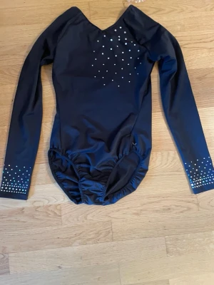 Svart gymnastikdräkt med strass - Svart långärmad gymnastikdräkt med V-ringning och silvriga strasstenar framtill samt på ärmsluten. Tillverkad i stretchigt material som ger bra rörelsefrihet, perfekt för gymnastik eller dans.