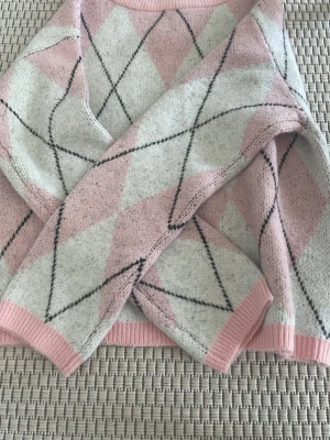 Rutig stickad tröja från 157, rosa/vit - Stickad tröja från 157 i mjukt material med klassiskt argylemönster i ljusrosa, vitt och svarta linjer. Tröjan har ribbade muddar och rund hals. Perfekt för dig som gillar en söt och trendig look.