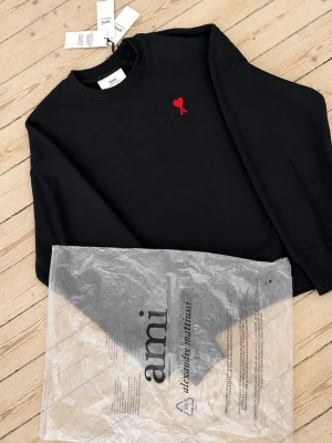 Svart sweatshirt från Ami Litet hjärta - Svart sweatshirt från Ami Alexandre Mattiussi med röd broderad hjärtdetalj på bröstet. Klassisk rund halsringning och långa ärmar. Tillverkad i mjuk bomull för en bekväm känsla. Perfekt för en stilren och avslappnad look. Size M-S Med påse och lappar.