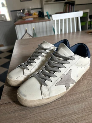 Vita sneakers med grå stjärna - Snygga vita sneakers från Golden Goose med grå mockadetaljer, inklusive en stor stjärna på sidan och matchande grå skosnören. Skorna har en låg silhuett och mörkblå hälkappa. Perfekta för en avslappnad och trendig stil.