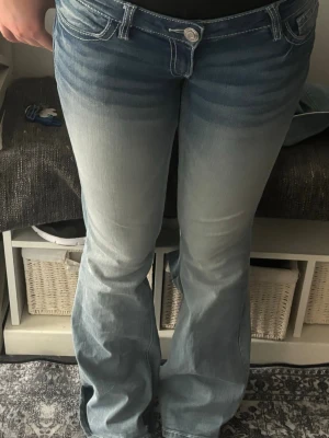 Ljusblå bootcut jeans med brodyr - Säljer ett par ljusblå bootcut jeans med snygg brodyr på bakfickan. Jeansen har låg midja, klassiska fem fickor och vita kontrastsömmar. Säljer då de tyvärr är för stora. Har en pytteliten reva. Kan även byta mot storlek S. Utöver revan perfekt skick