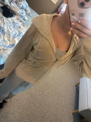 Beige velour hoodie med dragkedja - Snygg beige hoodie i mjuk velour med dragkedja framtill och fickor på sidorna. Hoodien har huva med snören och ribbade muddar vid ärmslut och nederkant. Perfekt för en chill och avslappnad look.