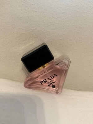Prada Virtual Flower parfym - Lyxig parfym från Prada med namnet Virtual Flower. Flaskan är trekantig i genomskinligt glas med ljusrosa innehåll och har en stor svart fyrkantig kork med gulddetalj. Perfekt för dig som gillar exklusiva dofter och snygg design på parfymflaskan.