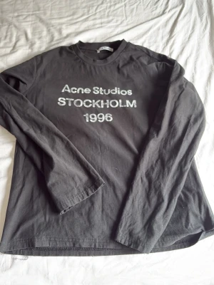 Svart långärmad topp Acne Studios - Svart långärmad topp från Acne Studios med vit text 'Acne Studios STOCKHOLM 1996' på bröstet. Klassisk rund halsringning och rak passform. Tillverkad i mjuk bomull, perfekt för en stilren och avslappnad look.