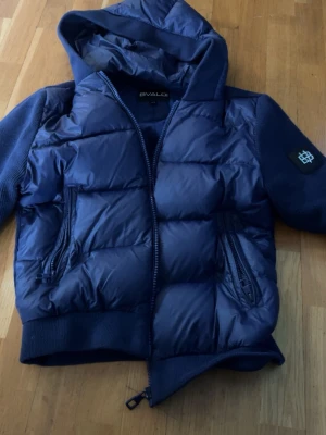Mörkblå cardigan med huva från Bvaldi - Snygg mörkblå cardigan från Svaldi med huva och dragkedja framtill. Jackan har två dragkedjeförsedda fickor och ribbade muddar. Ärmarna är i matchande tyg och det finns en patch på ena ärmen. Perfekt för lager-på-lager och streetwear.