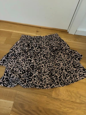 Leopardmönstrad volangkjol - Snygg kort volangkjol med leopardmönster i svart och rosa. Kjolen har resår i midjan och flera lager volanger som ger en cool och trendig look. Perfekt för dig som vill sticka ut lite extra.