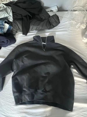 Svart halfzip hoodie - Svart hoodie med halfzip och hög krage. Tröjan har en enkel och clean design utan synliga loggor eller tryck. Tillverkad i mjukt sweatshirtmaterial och har breda muddar vid ärmslut och nederkant. Perfekt för en avslappnad och stilren look.