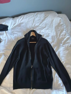 Svart ziptröja från Massimo Dutti - Stilren svart ziptröja från Massimo Dutti med hög krage och hel dragkedja. Tröjan är i ett mjukt stickat material och har en klassisk passform med långa ärmar. Perfekt för lager-på-lager och enkel att matcha med olika outfits. Nypris:900 kr