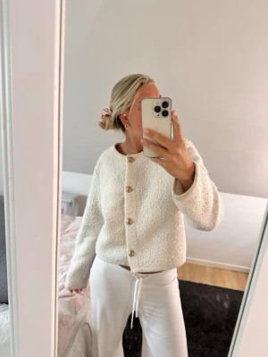 Vit kofta från Gina Tricot - Mysig vit kofta från Gina Tricot med rund halsringning och dekorativa guldknappar framtill. Koftan har en fluffig, bouclé-liknande struktur och raka ärmar. Perfekt för lager-på-lager och enkel att styla till olika outfits.