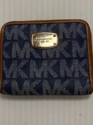 Blå MK plånbok i denim från Michael Kors - Snygg blå plånbok från Michael Kors i denim med bruna läderdetaljer och guldfärgad metallbricka framtill. Plånboken har ett tydligt MK-mönster över hela ytan och dragkedja runt. Perfekt för dig som gillar trendiga accessoarer med logga.