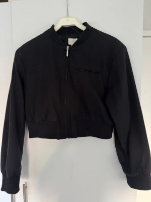 Svart cropped bomberjacka - Svart bomberjacka med croppad passform och ribbade muddar. Jackan har dragkedja framtill och en diskret bröstficka med dragkedja. Enkel och stilren design som passar till många outfits. Den är välanvänd och kan behöva noppras av lite