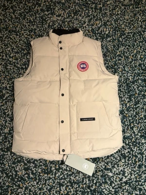 Beige dunväst från Canada Goose - Snygg dunväst från Canada Goose med klassisk logotyp på bröstet och svarta tryckknappar. Västen har två stora fickor framtill och hög krage. Perfekt för lager-på-lager och håller dig varm under kyliga dagar.