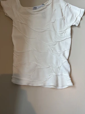 Vit strukturerad topp från Zara - Säljer en vit kortärmad topp från Zara med unik strukturerad yta och råa sömmar. Toppen har rund halsringning och är tillverkad i ett mjukt tyg som ger en cool och trendig look.