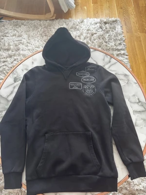 Svart hoodie från Mercier med patchar -  Köpt i England i jd sports, Sitter mer som en S eftersom den är typ slim fit Svart hoodie från Mercier med flera broderade patchar på bröstet. Klassisk känguruficka framtill, ribbade muddar och justerbar huva. Snygg streetstil med tydliga logodetaljer. Perfekt för dig som gillar en unik och trendig look.