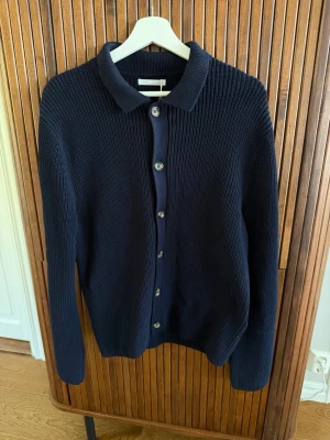 Mörkblå stickad kofta med krage - Mörkblå stickad kofta med klassisk krage och knäppning framtill. Koftan har långa ärmar och ribbstickad struktur som ger en stilren look. Perfekt att slänga på sig över en t-shirt eller skjorta.
