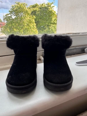 Svarta boots med fuskpäls - Säljer ett par svarta boots till barn med mjuk fuskpäls runt skaftet. Skorna har en rund tå och är tillverkade i mockaliknande material. Perfekta för kalla dagar när du vill hålla fötterna varma och snygga.