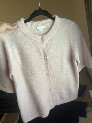 Ljusrosa kofta från Gina Tricot - Supersöt ljusrosa kofta från Gina Tricot med korta ärmar och pärlknappar framtill. Koftan är stickad i ett mjukt material och har ribbad kant vid halsen. Perfekt för en söt och trendig look.
