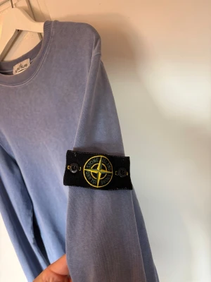 Ljusblå Stone Island sweatshirt - Ljusblå sweatshirt från Stone Island med klassisk rund halsringning och ikonisk kompasspatch på vänster ärm. Tröjan har en avslappnad passform och är tillverkad i mjuk bomull. Perfekt för en stilren och avslappnad look.