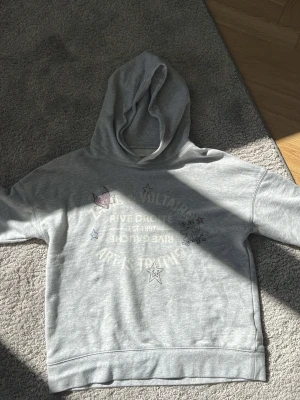 Grå hoodie från Zadig & Voltaire - Snygg ljusgrå hoodie från Zadig & Voltaire med tryck och stjärndetaljer på bröstet. Klassisk modell med huva och ribbade muddar. Kostar i nypris runt 1300 kr. Perfekt för en cool och trendig stil. Mjuk bomullsblandning som känns skön mot huden.