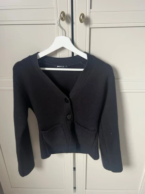 Svart kofta från Gina Tricot - Stilren svart kofta från Gina Tricot med två stora knappar framtill och praktiska fickor. Koftan har v-ringning, långa ärmar och är gjord i ett mjukt stickat material. Perfekt att slänga över en t-shirt eller topp för en enkel look.