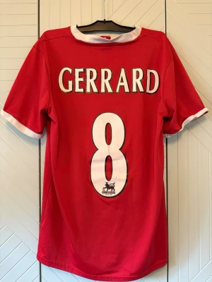 Liverpool Gerrard matchtröja röd - Röd Liverpool matchtröja med vita detaljer, Gerrard och nummer 8 på ryggen. Framsidan har Carlsberg-logga och klubbmärke. Klassisk design med korta ärmar och rund hals, perfekt för fotbollsfans.