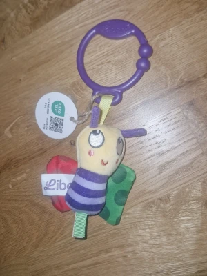 Libero mjuk bi-leksak med ring - Söt mjuk leksak från Libero i form av ett bi med lila och gula färger, gröna vingar med prickar och röd detalj. Fäst i en lila plast-ring för enkel upphängning, perfekt till barnvagn eller babyskydd. Vingarna är designade för att prassla vid beröring, och kroppen har olika mönster och färger.