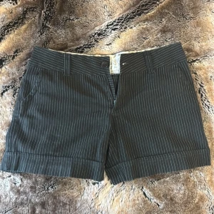 Svarta shorts med vita ränder - Snygga svarta shorts med tunna vita kritstrecksränder och uppvikta benslut. Lågmidjad passform med fickor fram och hällor för skärp. Insidan har ett mönstrat tyg med textdetaljer.  Tyvärr för små för mig