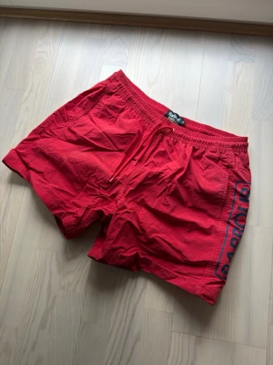 Röda badbyxor från Barbour - Snygga röda badbyxor från Barbour med elastisk midja och dragsko. Stora Barbour-loggan i blått på sidan, fickor fram och en bakficka som stängs. Perfekta för stranden eller poolen, tillverkade i lätt och snabbtorkande material. Är i storlek XL men passar verkligen L-M.