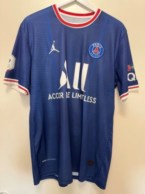 PSG Messi matchtröja blå - Blå Paris Saint-Germain matchtröja med Messi 30-tryck på ryggen. Tröjan har vita och röda detaljer vid krage och ärmslut, samt klubbmärke och sponsorloggor framtill. Tillverkad i lätt och ventilerande material, perfekt för fotbollsträning eller support.