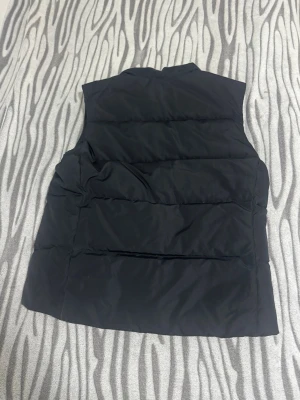 Svart dunväst från Champion - Snygg svart dunväst från Champion med klassisk quiltad design. Västen har hög krage och är tillverkad i ett glansigt material med dunfyllning som håller dig varm. Perfekt att slänga över en hoodie eller tröja för extra värme. 