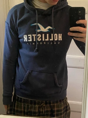 Mörkgrå Hollister hoodie med logga - Snygg mörkblå hoodie från Hollister med stor vit och blå logga på bröstet och texten 'California' under. Klassisk känguruficka framtill och justerbar huva med dragsko. Perfekt för chill dagar eller när du vill ha en avslappnad stil.