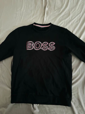 Svart sweatshirt från BOSS - Svart sweatshirt med rund hals och lång ärm från BOSS. Stor logotyp i vitt och rött framtill. Klassisk passform och ribbade muddar vid ärmslut och nederkant. Perfekt för en stilren och avslappnad look.