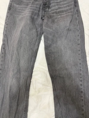 Grå raka jeans från 157 - Säljer ett par grå jeans från 157 med rak passform och klassisk femficksdesign. Jeansen har silverfärgade nitar och knappar samt normal midja. Materialet är denim i bomull och färgen är tvättad grå för en avslappnad look. Nytt skick