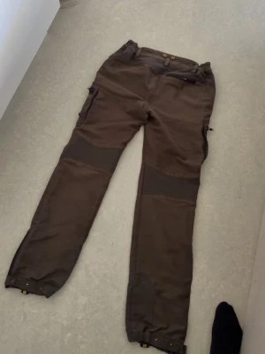 Bruna cargobyxor från Kayoba Outdoor - Snygga bruna cargobyxor från Kayoba Outdoor med förstärkta partier på knän och benslut. Byxorna har flera praktiska fickor och justerbara benslut, perfekta för äventyr utomhus. Materialet känns slitstarkt och är anpassat för outdoor.