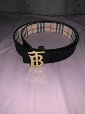 Svart Burberry bälte med guldspänne - Stilrent svart bälte från Burberry med klassiskt rutigt innerfoder och ett stort guldigt TB-spänne framtill. Bältet har en bred design och är tillverkat i skinn med subtila rutdetaljer på utsidan. Perfekt för att lyfta din outfit.
