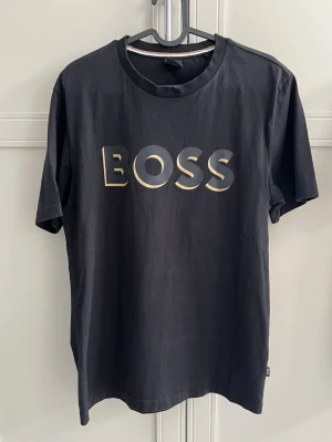 Svart BOSS t-shirt med tryck - Svart t-shirt från BOSS med stort guldfärgat BOSS-tryck på bröstet. Klassisk rund halsringning med snygg randig detalj på insidan av kragen. T-shirten har korta ärmar och är tillverkad i mjuk bomull för en bekväm känsla.