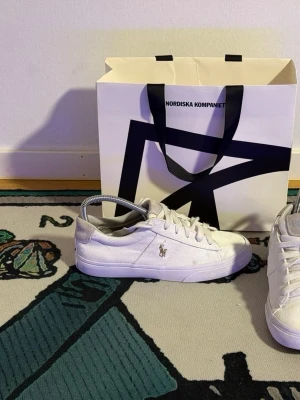 Vita Polo Ralph Lauren sneakers - Snygga vita sneakers från Polo Ralph Lauren med klassisk låg profil. Skorna har canvasmaterial, mockadetaljer vid hälen och den ikoniska Polo-loggan på sidan. Perfekta för en clean och stilren look. (Små i storleken kanske 36-37)