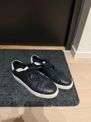 Marinblå sneakers från Gant Mc Julian - Stilrena marinblå sneakers från Gant med vita sulor och detaljer. Skorna har klassisk låg profil, rund tå och är tillverkade i läder med matchande marinblå skosnören. Perfekta för en clean och modern look.