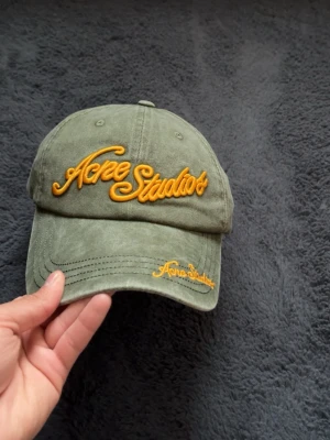 Grön keps från Acne Studios - Snygg grön keps från Acne Studios med gul broderad logga framtill och på skärmen. Klassisk böjd skärm och justerbart spänne bak. Tillverkad i bomullstyg med vintagekänsla. Perfekt accessoar för en avslappnad streetwear-look.