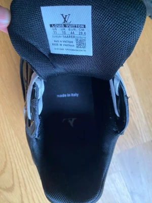 Louis Vuitton svarta sneakers - Svarta sneakers från Louis Vuitton med klassisk låg profil och diskret LV-logga på insidan. Skorna har svart ovandel i textil och syntetmaterial, samt svarta skosnören. Perfekta för dig som vill ha en stilren och exklusiv sneaker med lyxig känsla.