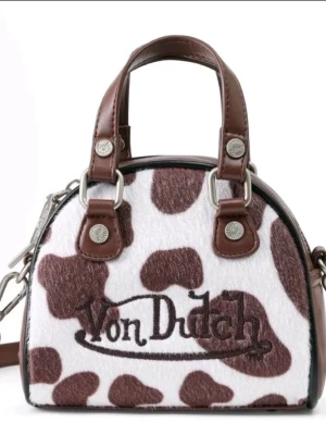 Von Dutch handväska med koskinnsmönster - Cool handväska från Von Dutch med brunt och vitt koskinnsmönster. Väskan har bruna läderdetaljer, silverfärgade metalldelar och broderad logga på framsidan. Perfekt för dig som vill sticka ut med en unik accessoar.
