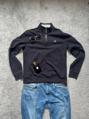 Morris half-zip  - Svart sweatshirt med half-zip ifrån Morris. Tröjan har ett broderat emblem på bröstet och ribbade muddar vid ärmslut och nederkant. Perfekt för en avslappnad och stilren look.