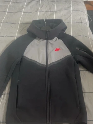 Svart och grå vindjacka från Nike - Nike vindjacka i svart och grått med röd logga på bröstet. Jackan har huva, hel dragkedja och sportig design. Perfekt för dig som gillar streetwear och vill ha en snygg och funktionell jacka till vardags. Knappt använd 